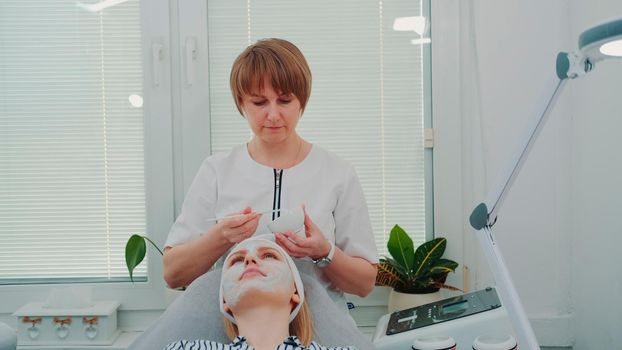 Beautician putting cream mask on woman's face at beauty salon. Facial skin care treatments.