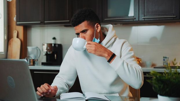Multiracial man with medical mask speaking by smartphone and working on computer at home kitchen. He also drinking coffee and making notes.
