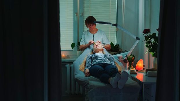 Beautician making relaxing facial massage with jade roller on woman's face at beauty salon with dim light around and pleasant atmosphere.