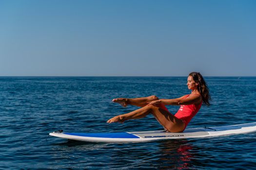 Sporty girl on a surfboard in the sea on a sunny summer day. In a red swimsuit, she sits in the splits on the sap. Summer entertainment on Stortom by the sea.