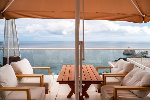 Restaurant with sea view. You can see rocks in the sea. Wooden tables and chairs with white cushions. Overcast, cloudy weather