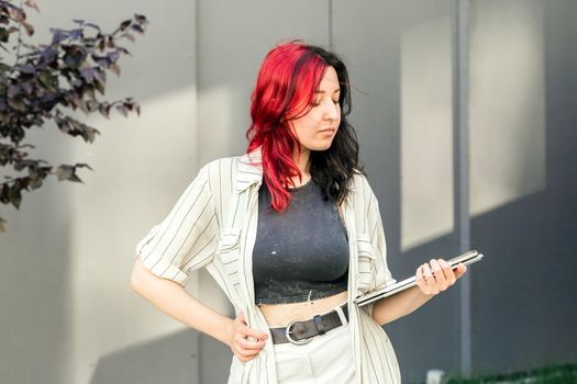 Portrait of a female student or businesswoman with creative red and black hair in smart casual clothing using digital tablet outdoors near the office building