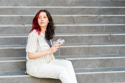 Young woman sitting on the stairs outdoors holding a silicone collapsible cup, reusable coffee-to-go tumbler.