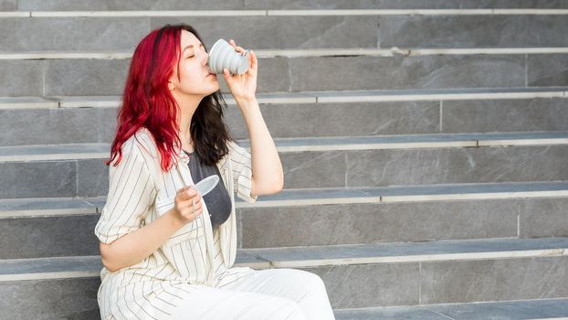 Young woman sitting on the stairs outdoors drinking from a silicone collapsible cup, reusable coffee-to-go tumbler.