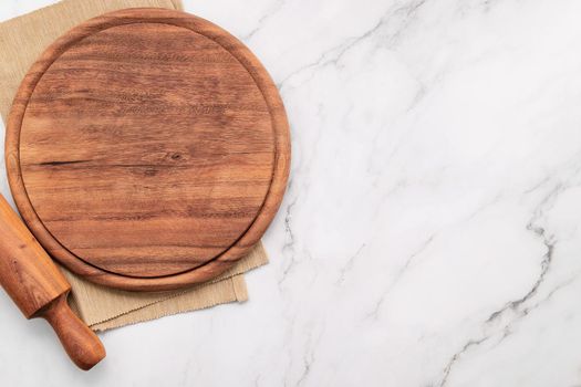 Empty wooden pizza platter with napkin and rolling pin set up on marble stone kitchen table. Pizza board and tablecloth on white marble background.