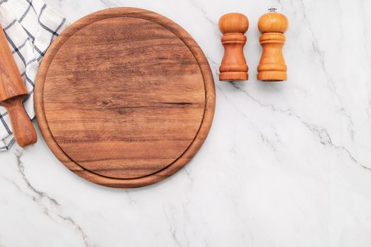 Empty wooden pizza platter with napkin and rolling pin set up on marble stone kitchen table. Pizza board and tablecloth on white marble background.