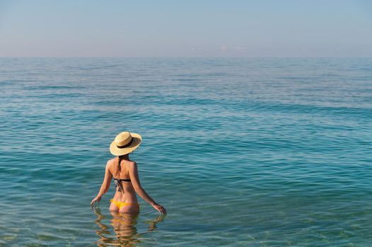Back view of a slender caucasian young woman standing waist-deep in calm transparent sea water in a swimsuit and a straw hat.
