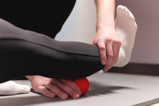 Young caucasian male masseur helping a female client to do myofascial massage of the calf muscle with a red massage ball. Close-up.