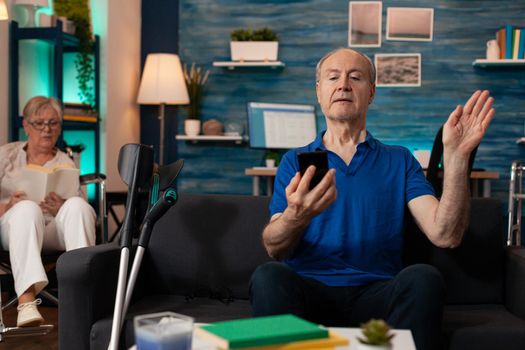 Elderly man using smartphone for video call communication in living room. Aged person waving at conference camera for online remote meeting. Woman sitting in wheelchair in background