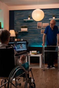 Disabled old woman holding laptop with video call talking to doctor and young girl in hospital ward. Invalid person in wheelchair using gadget for online remote conference at home