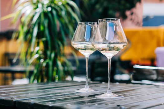 A wooden table in a restaurant with white wine. Wine glasses served for a party in a bar or a restaurant on terrace at a sunny day. Blurred background with plants. Still life scene