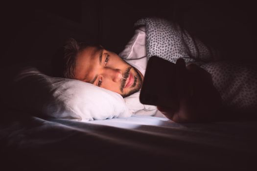 Young man lying in bed and using smartphone at night before sleeping. Social media addiction and insomnia. Communication and people concept . High quality photo