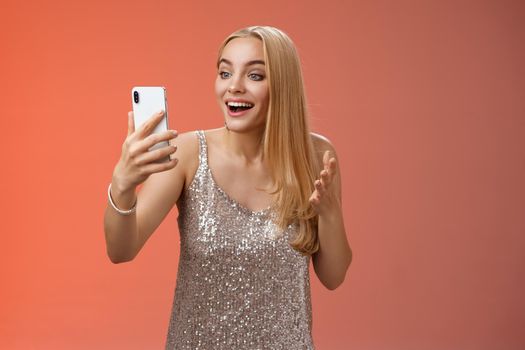 Charming elegant nice blond girl in silver dress talking video call speaking looking smartphone display amused surprised smiling happily have conversation sibling showing prom outfit.