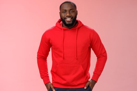 Handsome young happy black bearded guy white perfect smile hold hands pockets casually standing pink background talking have interesting nice conversation, wearing red hoodie. Copy space
