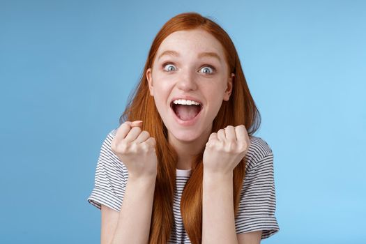 Yes awesome news great. Attractive surprised redhead girl cheering happy clench fists yelling yeah accomplish goal wide eyes excited celebrating win lottery triumphing joyfully, blue background.