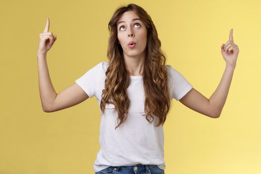 Impressed curious wondered attractive european woman long curly haircut look pointing index fingers up top copy space promo react astonished fascinated surprising event yellow background.