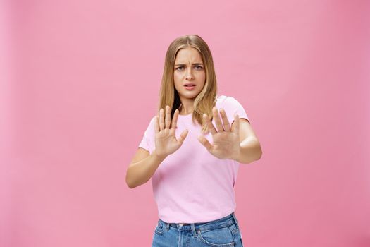 Stay away from me. Intense worried and displeased woman being victim of sexual harassment raising palms in defensive gesture making step backwards frowning asking stop, refusing over pink wall. Body language concept