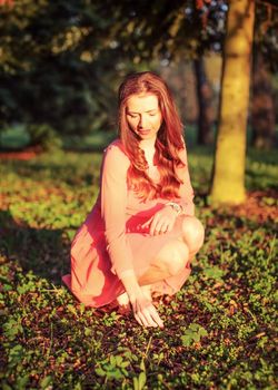 Young woman in pink dress crouching in the park, touching grass on ground, during evening sunset light.