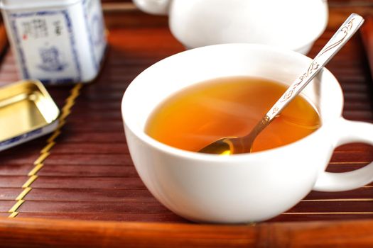 White porcelain cup of hot steaming tea, with spoon, placed on bamboo tray.