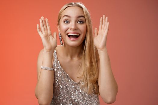 Impressed charming entertained blond european 25s woman in stylish silver glamour dress having fun playing peekaboo smiling joyfully hold palms near face laugh amused, standing red background.