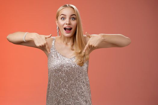 Impressed excited attractive glamour blond girl in silver glittering dress gasping thrilled pointing down glance camera fascinated check out fabulous awesome promo, standing amused red background.