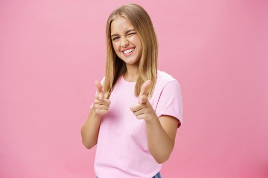 Woman expressing positive attitude towards camera pointing with fingers and winking joyfully smiling being uplifted, standing in good mood with optimistic gestures against pink background. Copy space