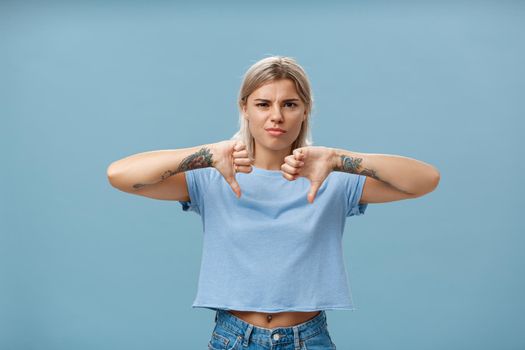 No way mate dislike. Portrait of dissatisfied bossy female tattoo artist with tattoos on arms frowning from displeasure showing thumbs down in disapproval standing over blue background. Copy space