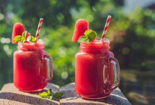 Healthy watermelon smoothie in Mason jars with mint and striped straws against the background of greenery.