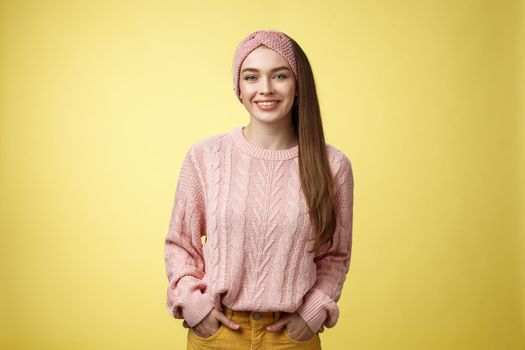Charming friendly young girl in knitted sweater and headband holding hands in pockets, smiling, looking positive motivated achieving goals having great day, posing positive against yellow background.