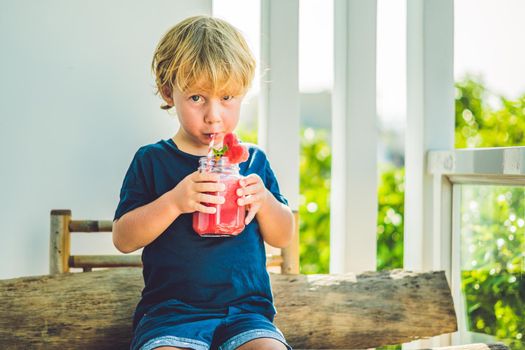 The boy is holding Healthy watermelon smoothie in Mason jar with mint and striped straws on a wood background.