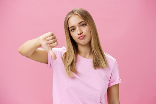 Displeased irritated and moody european woman with fair hair pursing lips annoyed showing thumbs down in dislike standing pissed and unimpressed giving negative feedback against pink wall. Lifestyle.