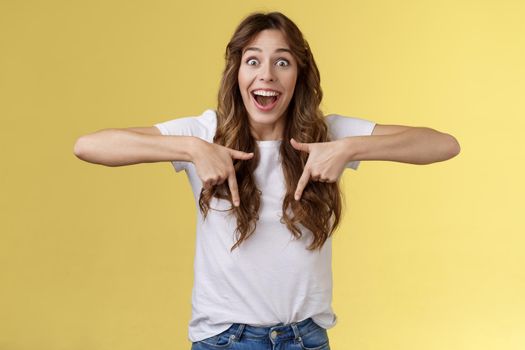 Excited happy cheerful impressed cute brunette girl long haircut smiling broadly surprised react pleased pointing down share excellent positive news stand joyfully yellow background promoting.