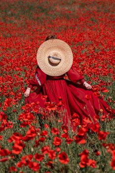 Young woman stands with her back in a long red dress and hat, posing on a large field of red poppies.