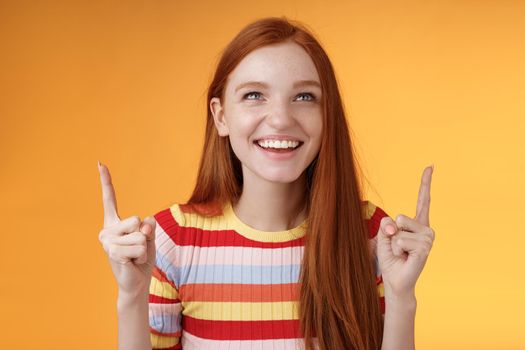 Happy cheerful redhead girl having fun amusement park laughing joyfully pointing look up index fingers upwards enjoy entertainment standing orange background amused grinning joyfully.