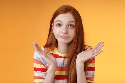 Clueless unbothered young redhead silly european girl 20s shrugging hands spread sideways smirking sorry cannot answer standing unaware confused puzzled give reply, orange background.