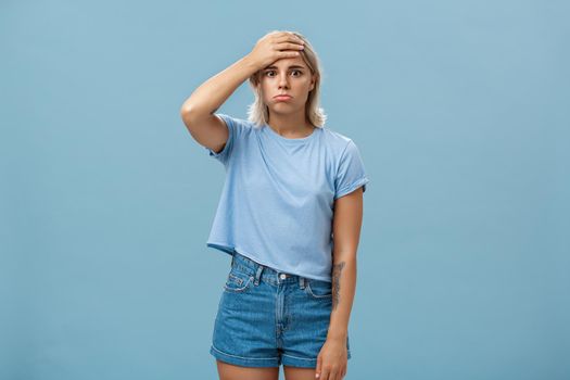 Lifestyle. Concerned and troubled young teenage girl being shocked with loads of responsibilities holding hand on forehead staring spaced out and pouting from tiresome perplexed situation over blue wall.