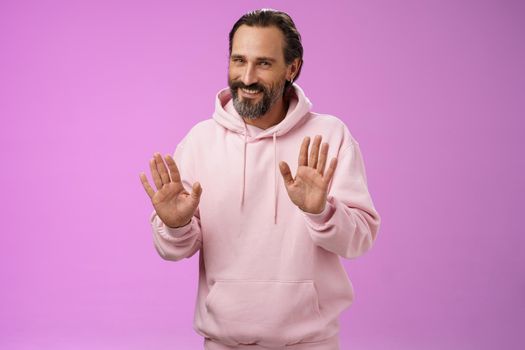 No thanks pass. Charming polite manly adult bearded man grey hair in pink hoodie apologizing refusal raise palms rejection gesture declining offer smiling say sorry not interested, purple background.
