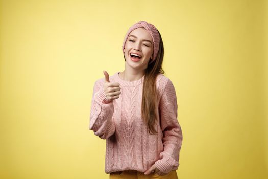 Happy amused beautiful young european girl in sweater, knitted headband showing good gesture, satisfied with service provided, winking sassy smiling with thumb-up in approval, giving recommendation.