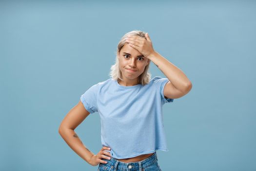 Lifestyle. Girl cannot deal with naughty kids being left alone while mom on shopping holding hand on forehead breathing out losing temper being in troublesome perplexed situation tired over blue background.