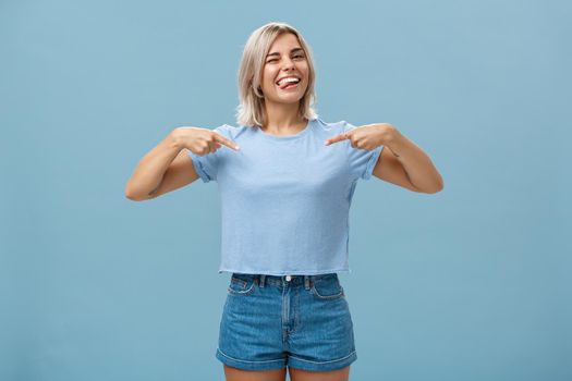 Yeah proud of myself. Self-assured joyful and carefree attractive blonde female with tanned skin and tattoos pointing at herself winking and sticking out tongue with broad smile over blue wall.