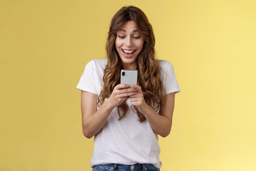 Excited cheerful happy curly-haired silly girl receive surprising good news hold smartphone smiling delighted look mobile phone screen answering b-day invitation stand yellow background upbeat. Lifestyle.