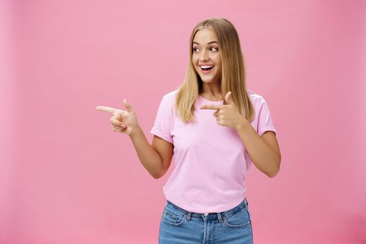 Happy cheerful young chubby girl with fair hair pointing and looking left with pleased excited smile turning attention to interesting copy space against pink background, expressing positive emotions.