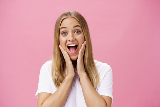 Portrait of amused delighted friendly and attractive european woman with fair hair touching cheeks from amusement and surprise smiling broadly gazing with admiration at camera over pink background. Emotions concept