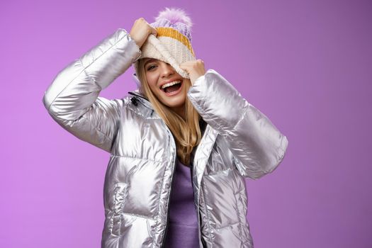 Amused carefree happy smiling playful attractive woman having fun laughing joyfully pulling hat face hiding peeking one eye camera grinning chuckling fool around have fun, purple background.