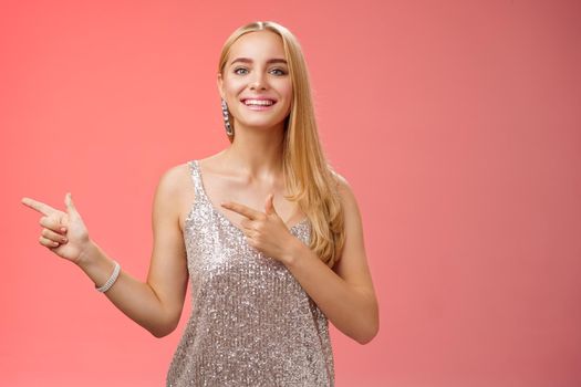 Proud happy good-looking stylish blond attractive caucasian woman in silver luxurious glamour dress pointing left smiling broadly showing friends new apartment, standing red background.