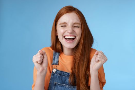 Lifestyle. Sincere happy rejocing ginger girl close eyes smiling broadly say yes waving clenched fists joyfully celebrate enterting university dream come true winning prize triumphing cheerfully blue background.