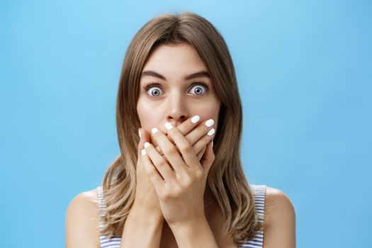 Close-up shot of shocked speechless woman covering mouth with pressed palms popping eyes at camera from surprise and worry, gasping witnessing terrible scene, feeling scared and intense over blue wall.