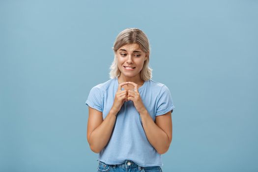 Lifestyle. Studio shot of insecure and timid silly girlfriend with blond hair clenching teeth looking shy down and steepling index fingers unconfident trying ask out boy she likes over blue background.