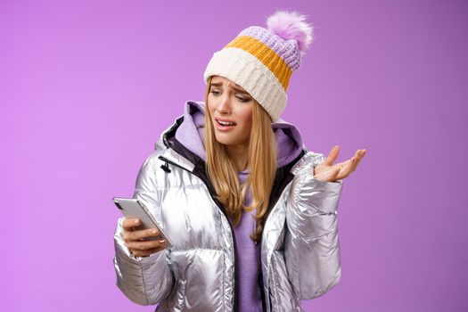 Upset disappointed attractive whining gloomy blond girl in silver jacket standing outside hat holding smartphone shrugging raising hand dismay complaining slow mobile internet, purple background.
