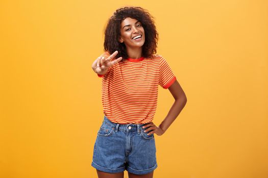 Friendly carefree and happy good-looking tall african american female model with curly hairstyle holding hand on hip tilting head and smiling joyfully showing peace gesture over orange wall. Copy space